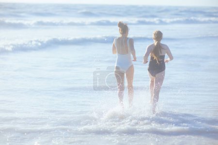 Foto de Visto desde atrás elegante madre e hija adolescente en la playa en traje de baño corriendo. - Imagen libre de derechos