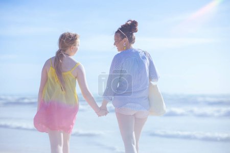 Foto de Visto desde atrás elegante madre e hija adolescente en la playa caminando. - Imagen libre de derechos