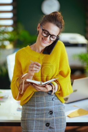 Photo for Happy 40 years old small business owner woman in yellow sweater with notebook talking on a smartphone in the modern green office. - Royalty Free Image