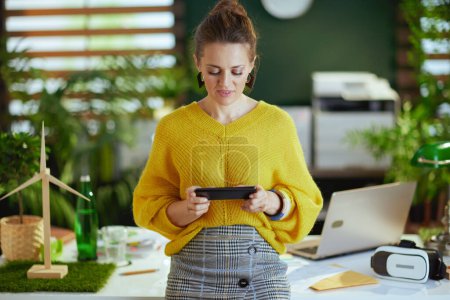 Téléchargez les photos : Jeune propriétaire de petite entreprise femme en pull jaune dans le bureau vert moderne en utilisant smartphone. - en image libre de droit