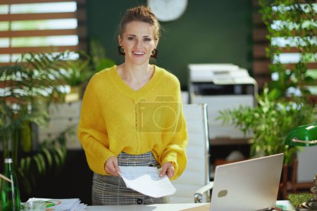 Téléchargez les photos : Heureuse femme propriétaire de petite entreprise moderne en pull jaune avec ordinateur portable travaillant avec des documents dans le bureau vert moderne. - en image libre de droit
