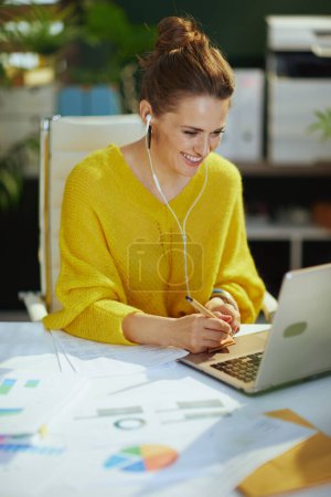 Foto de Sonriente de moda de mediana edad dueño de una pequeña empresa mujer en suéter amarillo con portátil hablando videollamada en la moderna oficina verde. - Imagen libre de derechos