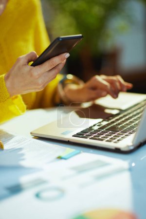 Foto de Primer plano de la mujer propietaria de una pequeña empresa en suéter amarillo con teléfono inteligente y portátil en la oficina verde. - Imagen libre de derechos