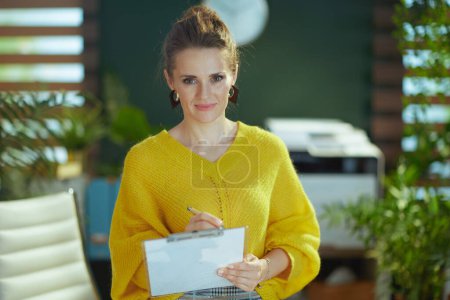 Téléchargez les photos : Élégante propriétaire de petite entreprise femme en pull jaune avec presse-papiers dans le bureau vert moderne. - en image libre de droit