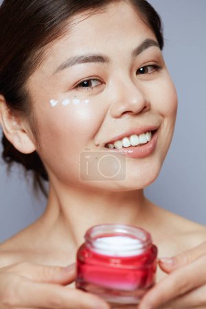 Téléchargez les photos : Portrait de femme asiatique moderne avec pot de crème faciale et crème pour les yeux sur le visage sur fond bleu. - en image libre de droit