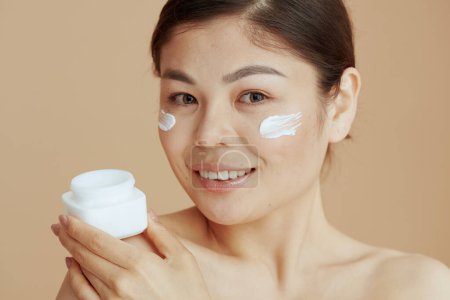 Foto de Retrato de mujer moderna con frasco de crema facial y crema facial en la cara aislada sobre fondo beige. - Imagen libre de derechos
