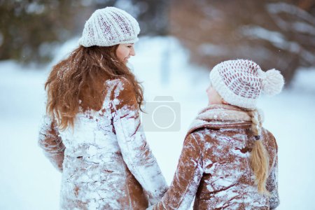 Foto de Smiling modern mother and daughter in coat, hat, scarf and mittens in snowy clothes walking outdoors in the city park in winter. - Imagen libre de derechos