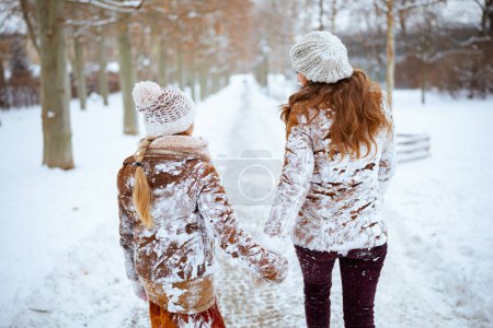 Foto de Seen from behind elegant mother and child in coat, hat, scarf and mittens in snowy clothes walking outdoors in the city park in winter. - Imagen libre de derechos