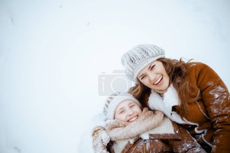 Foto de Sonriendo elegante madre e hijo en abrigo, sombrero, bufanda y manoplas que yacen en la nieve al aire libre en el parque de la ciudad en invierno. - Imagen libre de derechos