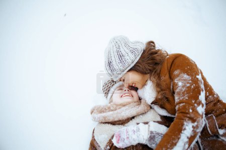 Foto de Happy modern mother and daughter in coat, hat, scarf and mittens laying in snow outdoors in the city park in winter. - Imagen libre de derechos