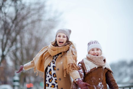 Foto de Smiling elegant mother and daughter in coat, hat, scarf and mittens catching snow outdoors in the city in winter. - Imagen libre de derechos