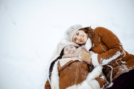 Photo for Smiling modern mother and child in coat, hat, scarf and mittens laying in snow outdoors in the city park in winter. - Royalty Free Image