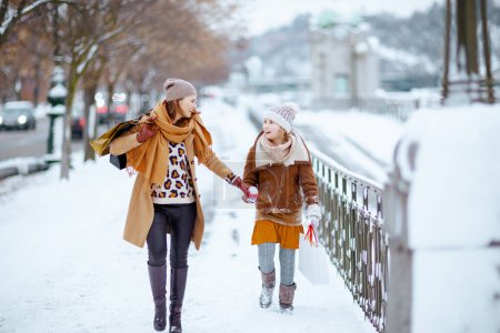 Photo for Happy elegant mother and child in coat, hat, scarf and mittens with shopping bags walking outdoors in the city in winter. - Royalty Free Image