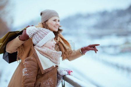 Foto de Happy stylish mother and daughter in coat, hat, scarf and mittens with shopping bags pointing at something outdoors in the city in winter. - Imagen libre de derechos