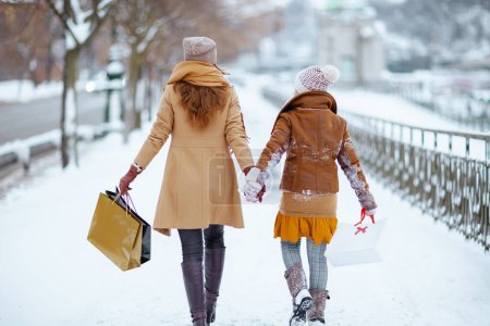Foto de Seen from behind mother and child in coat, hat, scarf and mittens with shopping bags walking outdoors in the city in winter. - Imagen libre de derechos