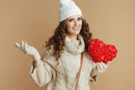 Photo for Hello winter. smiling elegant middle aged woman in beige sweater, mittens and hat against beige background with red heart. - Royalty Free Image
