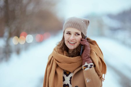 Foto de Happy elegant female in brown hat and scarf in camel coat with gloves using a smartphone outside in the city in winter. - Imagen libre de derechos