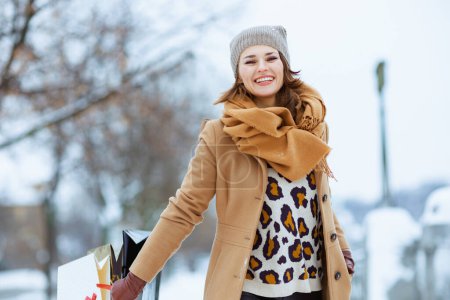 Téléchargez les photos : Happy elegant female in brown hat and scarf in camel coat with shopping bags outside in the city in winter. - en image libre de droit