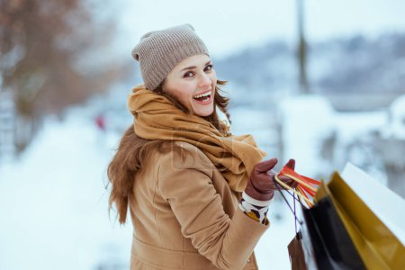 Foto de Smiling modern 40 years old woman in brown hat and scarf in camel coat with gloves and shopping bags outside in the city in winter. - Imagen libre de derechos