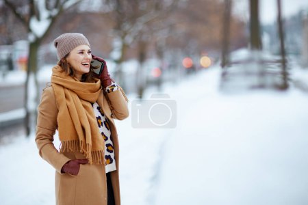Téléchargez les photos : Smiling stylish female in brown hat and scarf in camel coat with gloves using a smartphone outside in the city in winter. - en image libre de droit