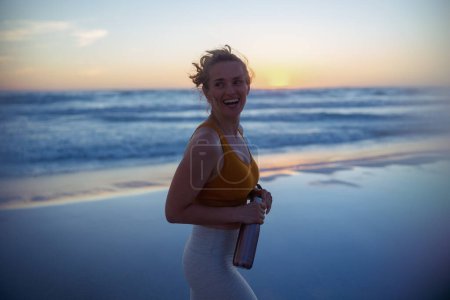Foto de Happy active woman jogger in fitness clothes with bottle of water walking at the beach at sunset. - Imagen libre de derechos