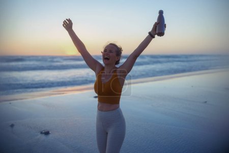 Téléchargez les photos : Happy fit sports woman in fitness clothes with bottle of water rejoicing at the beach at sunset. - en image libre de droit