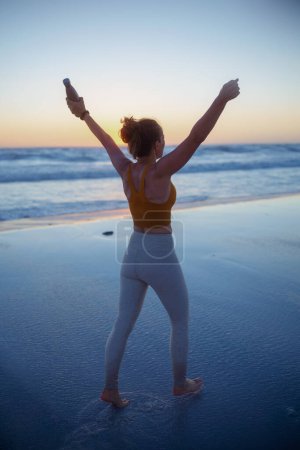 Téléchargez les photos : Seen from behind fit sports woman in fitness clothes with bottle of water rejoicing at the beach at sundown. - en image libre de droit