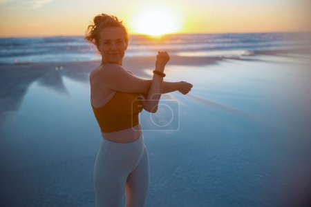 Foto de Happy healthy woman jogger in fitness clothes at the beach in the evening stretching. - Imagen libre de derechos