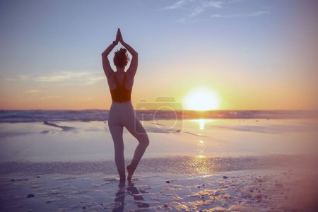 Foto de Seen from behind fitness woman jogger in fitness clothes at the beach at sunset meditating. - Imagen libre de derechos