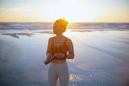 Téléchargez les photos : Relaxed active sports woman in sport clothes with bottle of water at the beach at sunset. - en image libre de droit