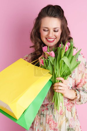 Foto de Happy young woman with long wavy brunette hair with tulips bouquet and shopping bags isolated on pink background. - Imagen libre de derechos