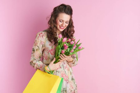 Téléchargez les photos : Happy modern woman in floral dress with tulips bouquet and shopping bags isolated on pink. - en image libre de droit