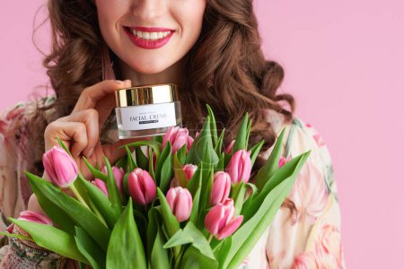 Foto de Closeup on smiling female with long wavy brunette hair with tulips bouquet and cosmetic jar against pink background. - Imagen libre de derechos