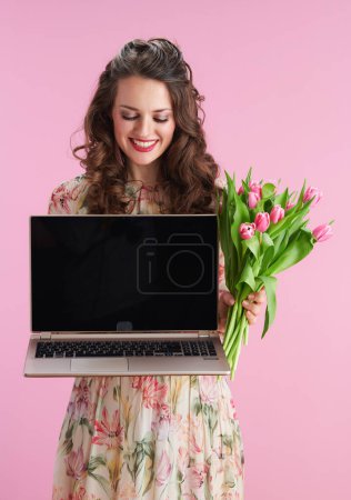 Foto de Smiling middle aged woman in floral dress with tulips bouquet showing laptop blank screen against pink background. - Imagen libre de derechos