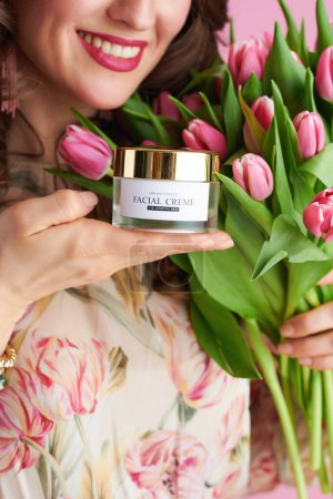 Foto de Closeup on smiling woman with tulips bouquet and cosmetic jar isolated on pink. - Imagen libre de derechos