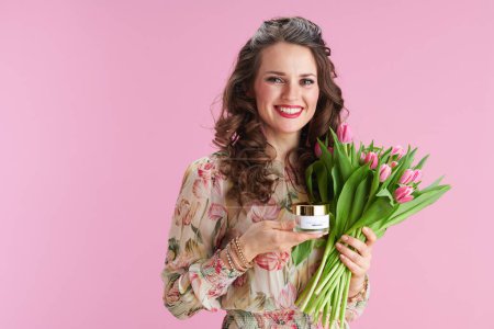 Portrait of happy trendy 40 years old woman with long wavy brunette hair with tulips bouquet and cosmetic jar against pink background.