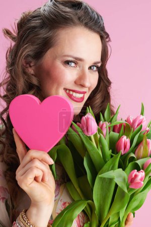 Téléchargez les photos : Heureuse élégante femme d'âge moyen avec de longs cheveux bruns ondulés avec bouquet de tulipes et coeur rose sur fond rose. - en image libre de droit