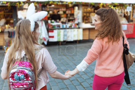 Foto de Easter fun. Seen from behind young mother and teenage daughter with easter bunny ears walking at the fair in the city. - Imagen libre de derechos