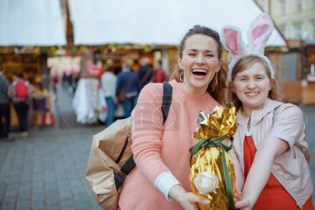 Foto de Easter fun. Portrait of smiling young mother and child with golden easter egg at the fair in the city. - Imagen libre de derechos