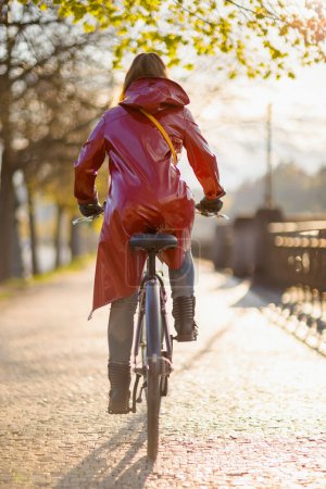 Foto de Visto desde atrás mujer con abrigo de lluvia rojo afuera en la ciudad montando en bicicleta. - Imagen libre de derechos
