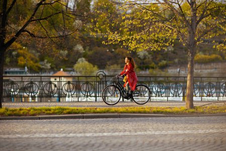 Photo for Modern middle aged woman in red rain coat outdoors on the city street riding bicycle. - Royalty Free Image