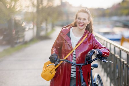 Photo for Smiling modern woman in red rain coat with bicycle outside on the city street. - Royalty Free Image