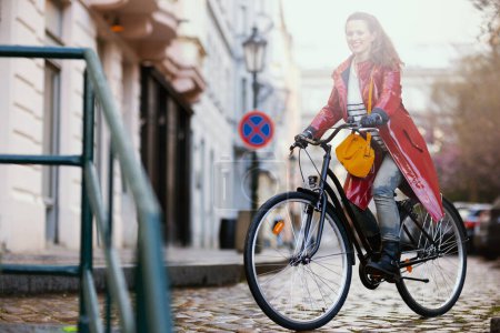 glücklich stylische 40-jährige Frau in rotem Regenmantel im Freien auf der Stadtstraße mit dem Fahrrad.
