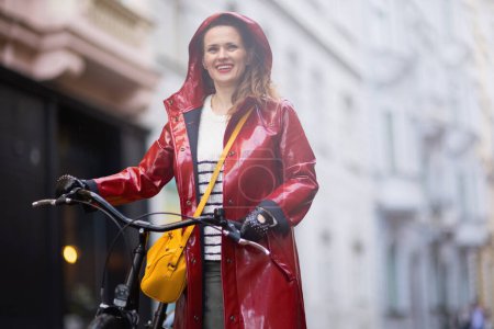 Téléchargez les photos : Smiling elegant female in red rain coat with bicycle in the rain outside on the city street. - en image libre de droit