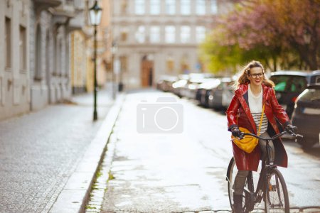 Téléchargez les photos : Femme élégante souriante en imperméable rouge à l'extérieur sur la rue de la ville à vélo. - en image libre de droit