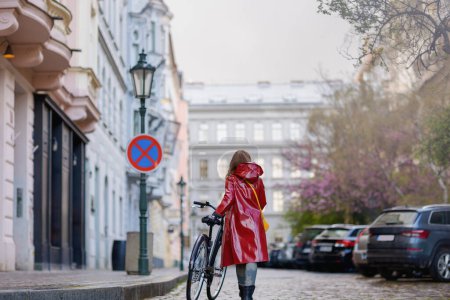 Foto de Seen from behind woman in red rain coat with bicycle walking in the rain outdoors on the city street. - Imagen libre de derechos