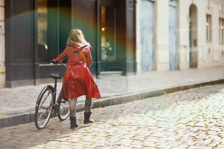 Foto de Seen from behind woman in red rain coat with bicycle walking outside on the city street. - Imagen libre de derechos