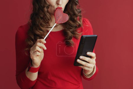 Photo for Happy Valentine. Closeup on middle aged woman isolated on red background with heart shaped candy on stick using smartphone applications. - Royalty Free Image