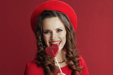 Photo for Happy Valentine. smiling elegant 40 years old woman in red dress and beret against red background with heart shaped candy on stick. - Royalty Free Image