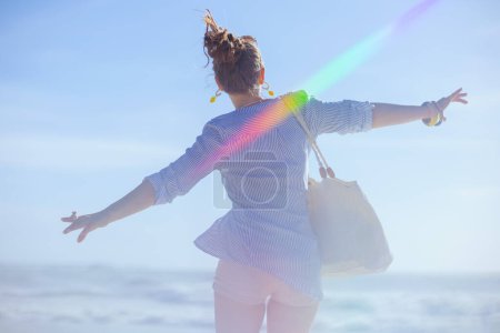 Téléchargez les photos : Vu de derrière femme d'âge moyen avec sac de paille blanche se réjouissant à la plage. - en image libre de droit